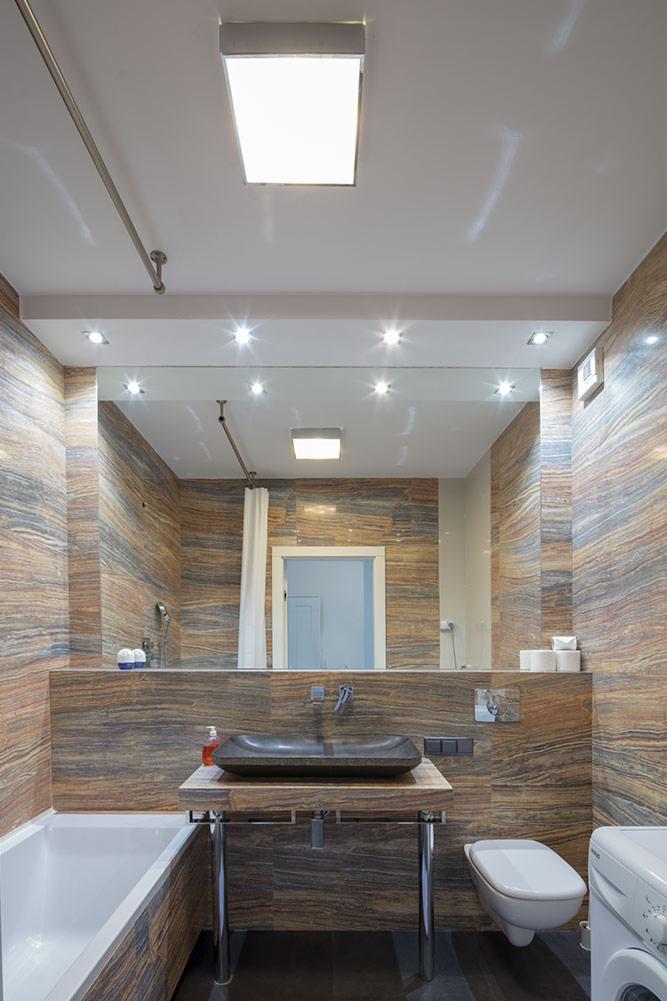 Nowoczesna łazienka z kamienną umywalką na chromowanym stelażu, płytki imitujące drewno, oświetlenie punktowe i biała armatura.