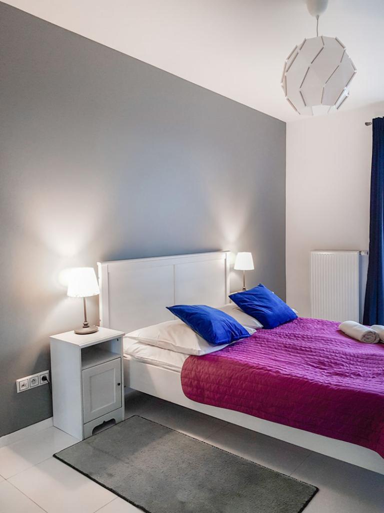 Pokój z łóżkiem małżeńskim, fioletową narzutą, niebieskimi poduszkami i szafką nocną z lampką, szary dywanik na białej podłodze.