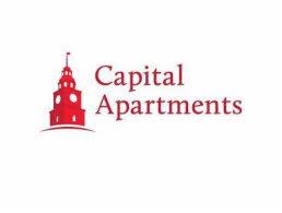 Czerwone logo Capital Apartments przedstawiające stylizowaną wieżę zegarową.