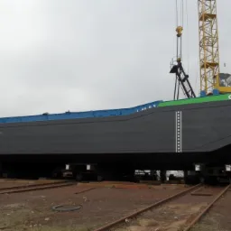 Barka 'G 502' po renowacji na torach, z widocznymi dźwigami w tle, świeżo pomalowana na szaro z zielonym pasem i niebieskim wykończeniem, z adresem strony internetowej na burcie.