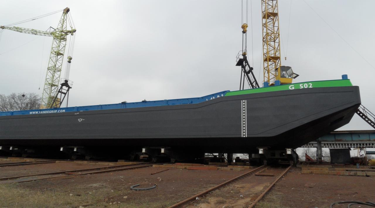 Barka 'G 502' po renowacji na torach, z widocznymi dźwigami w tle, świeżo pomalowana na szaro z zielonym pasem i niebieskim wykończeniem, z adresem strony internetowej na burcie.