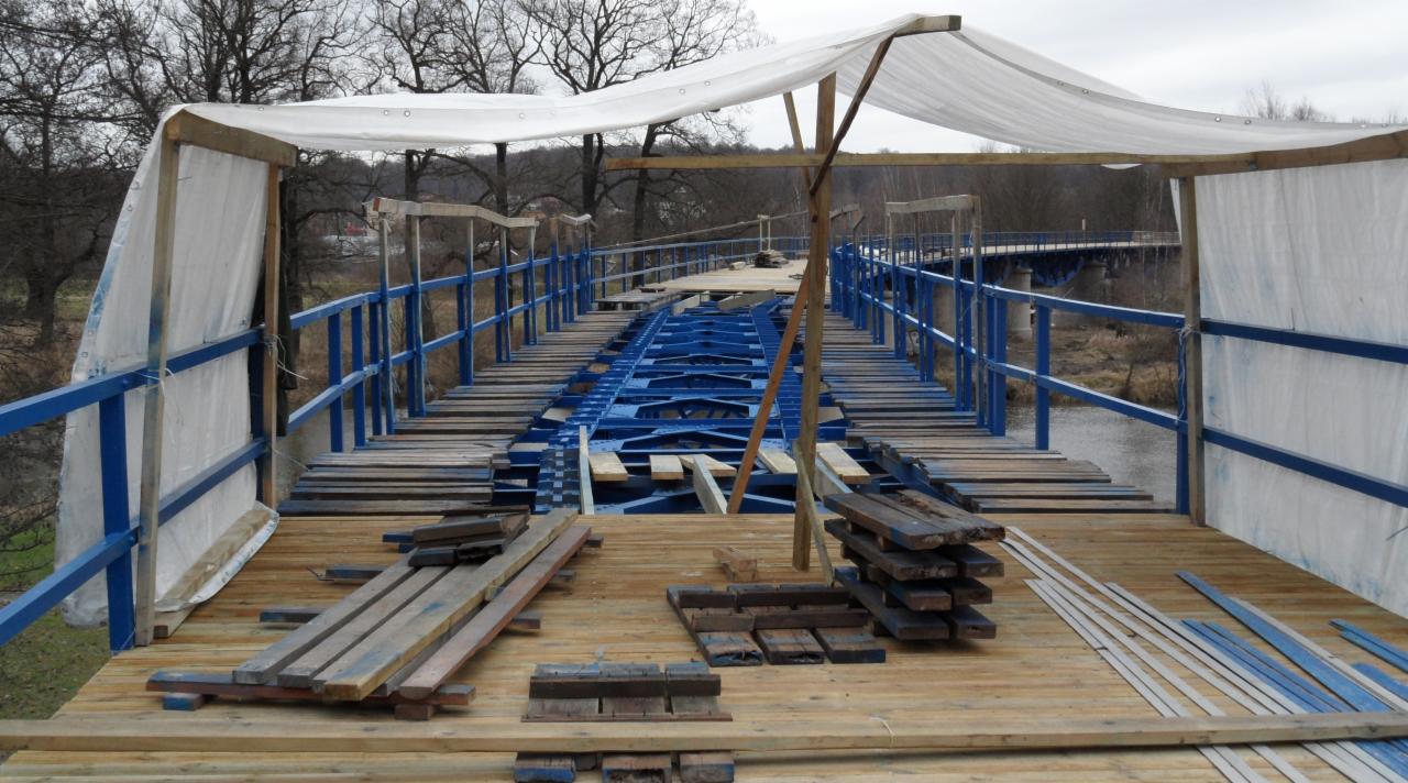 Most w trakcie renowacji: pomalowana na niebiesko metalowa konstrukcja, drewniane deski podłoża, częściowo osłonięty białą plandeką, w tle widać drugi most.