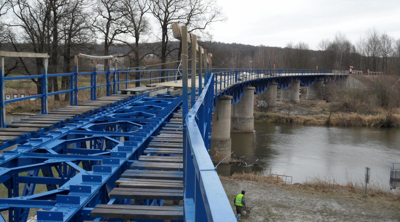 Niebieski most nad rzeką w trakcie renowacji, widoczne drewniane elementy pomostu i betonowe filary. Pracownik w odblaskowej kamizelce z taczką na dole zdjęcia.