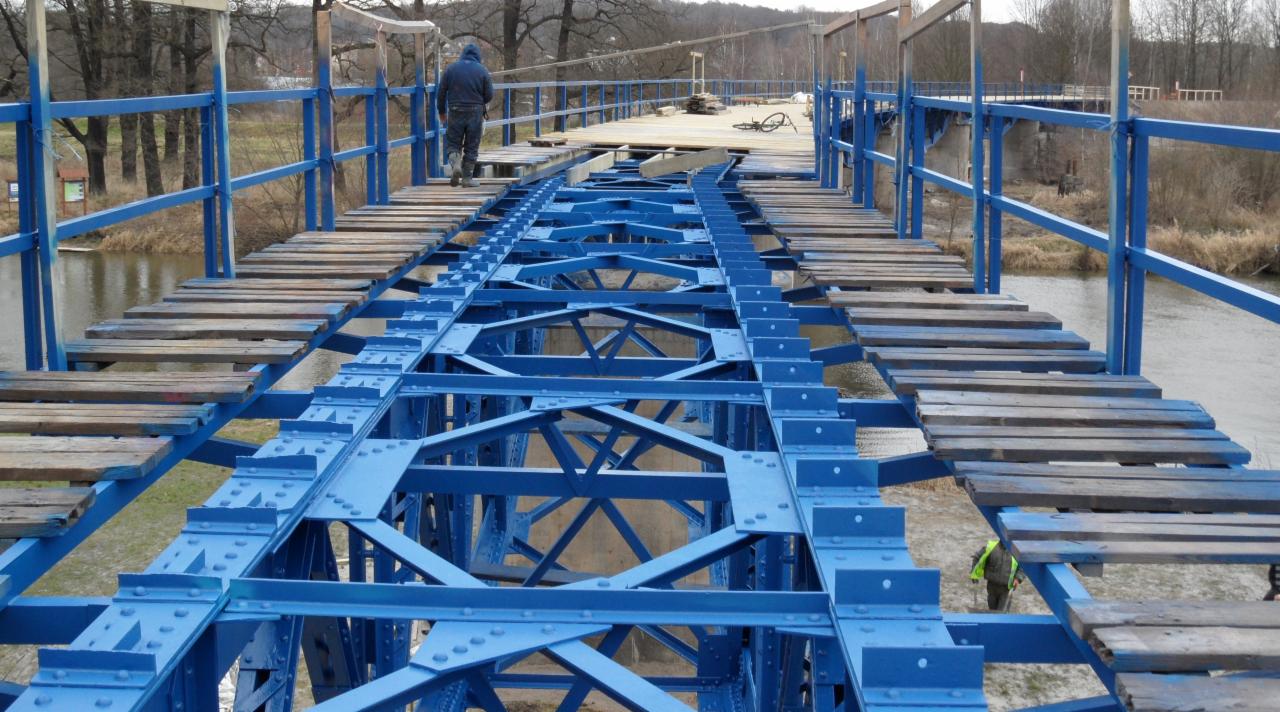 Niebieska konstrukcja stalowa mostu w trakcie renowacji, z częściowo ułożonymi drewnianymi deskami pomostu; w tle rzeka i drzewa.