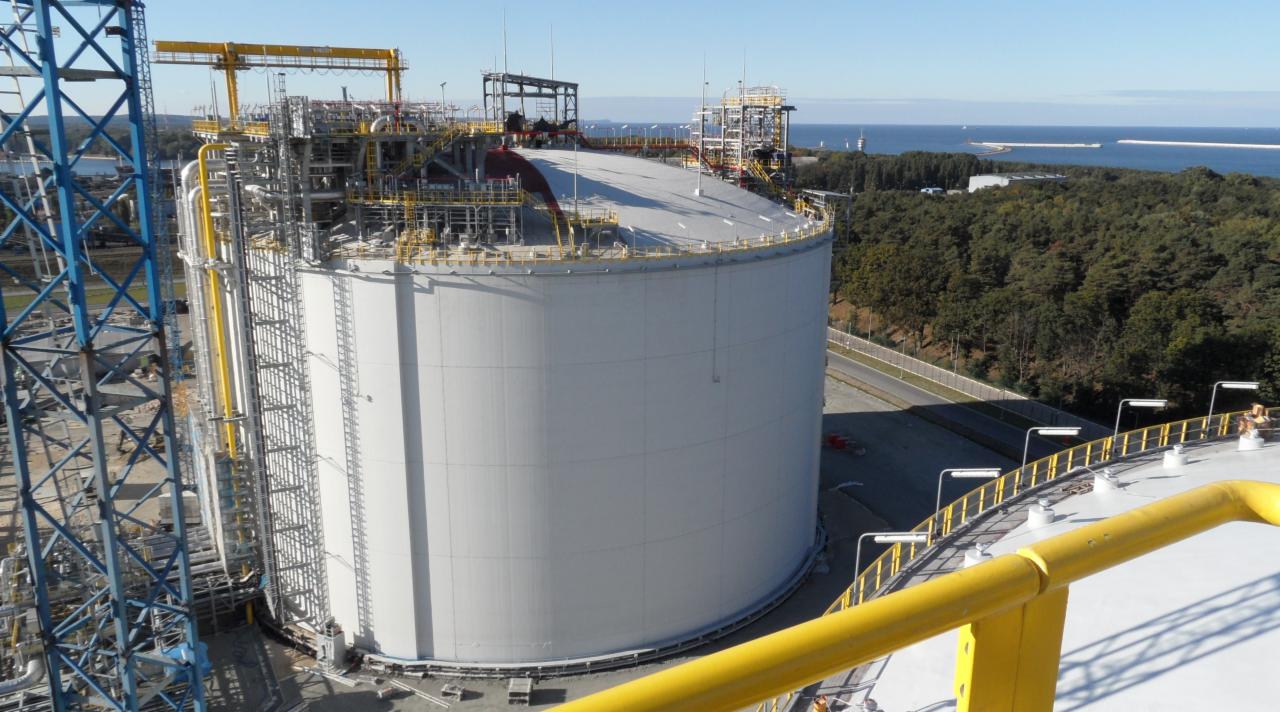 Cylindryczny zbiornik przemysłowy z instalacjami, widoczny z góry, z lasem i morzem w tle.