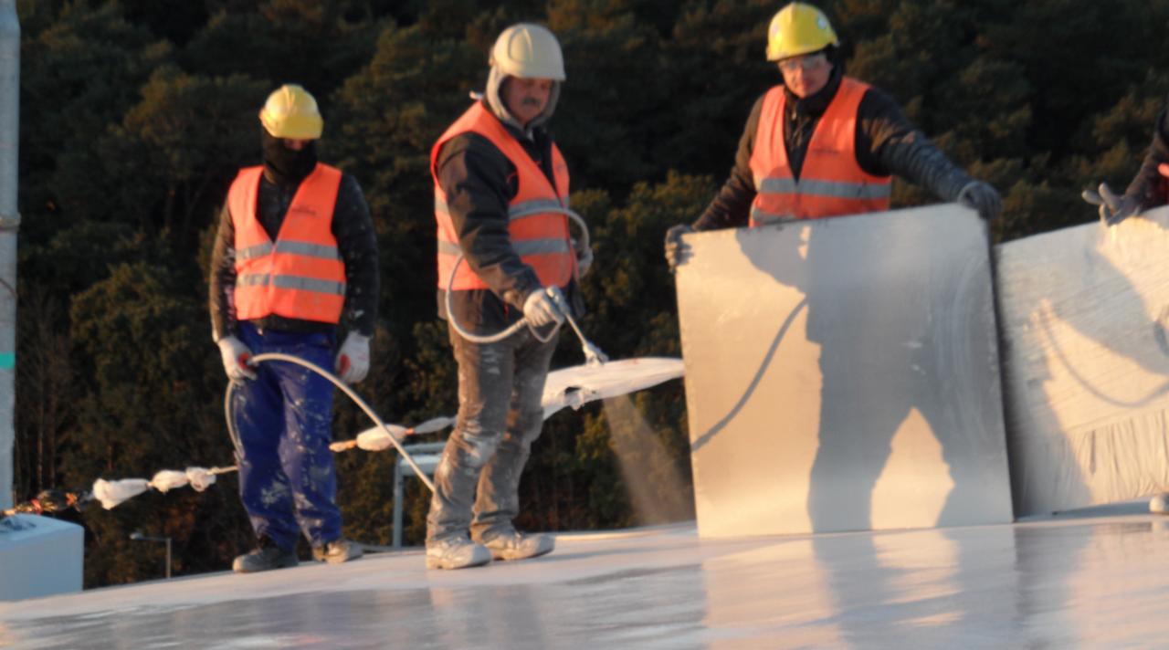 Pracownicy w kaskach i kamizelkach ochronnych nanoszą białą powłokę natryskową na płaską powierzchnię dachu, widoczny wąż i pistolet natryskowy, w tle zieleń drzew.