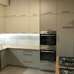 Nowoczesna kuchnia w odcieniach szarości z drewnianym blatem, zabudowane piekarniki Electrolux, płyta gazowa i okap, widoczne szafki górne i dolne z podświetleniem LED.