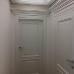 Plichta sławomir - Dwa białe, klasyczne drzwi wewnętrzne z dekoracyjnymi listwami i srebrnymi klamkami, widoczne w korytarzu z drewnianą podłogą i sufitem z oświetleniem, zabezpieczonym niebieską taśmą malarską.