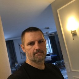 Plichta sławomir - Mężczyzna w czarnej koszulce z logo firmy DDD na tle wnętrza mieszkania z widoczną lampą ścienną i fragmentem salonu.