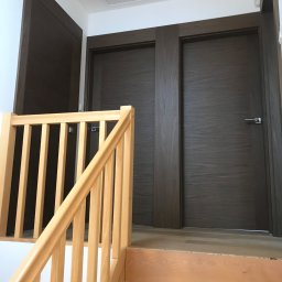 Plichta sławomir - Dwa ciemne, drewniane drzwi z klamkami, widoczne zza jasnych, drewnianych schodów z barierką w nowym domu.
