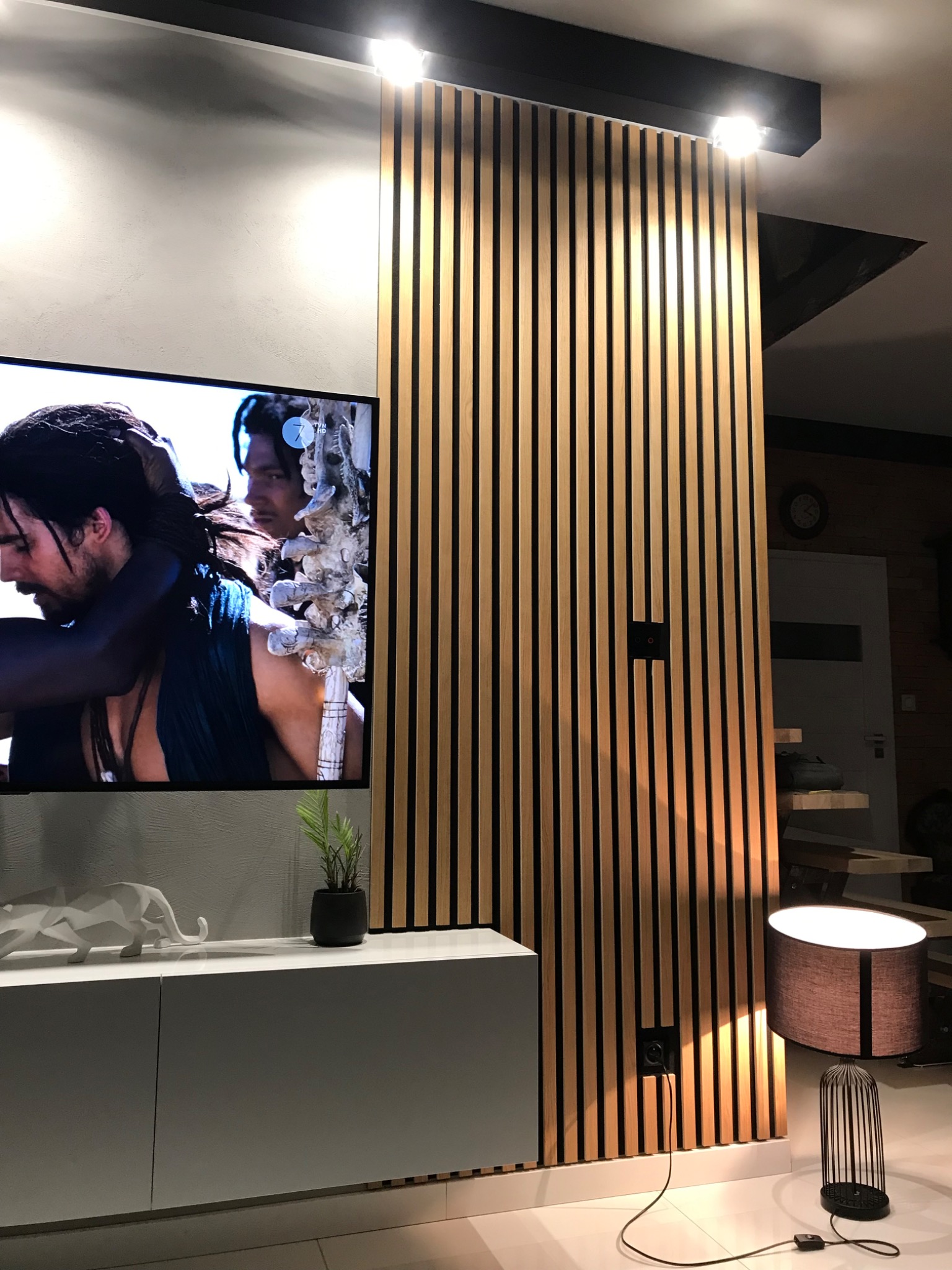 Nowoczesna aranżacja wnętrza z drewnianą ścianką działową z pionowych listew, telewizorem z włączonym filmem, białą szafką RTV z dekoracjami i lampą podłogową, tworząca przytulną przestrzeń.