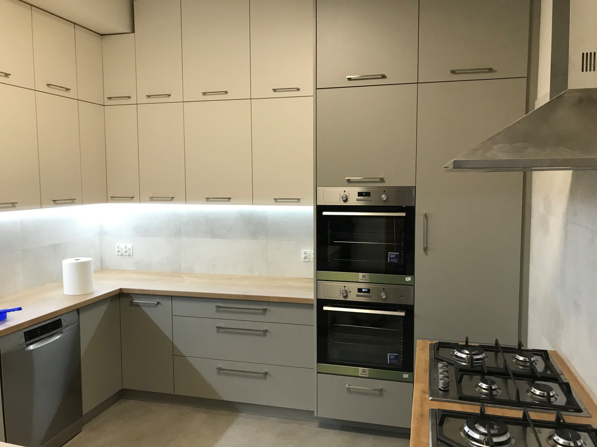 Nowoczesna kuchnia w odcieniach szarości z drewnianym blatem, zabudowane piekarniki Electrolux, płyta gazowa i okap, widoczne szafki górne i dolne z podświetleniem LED.