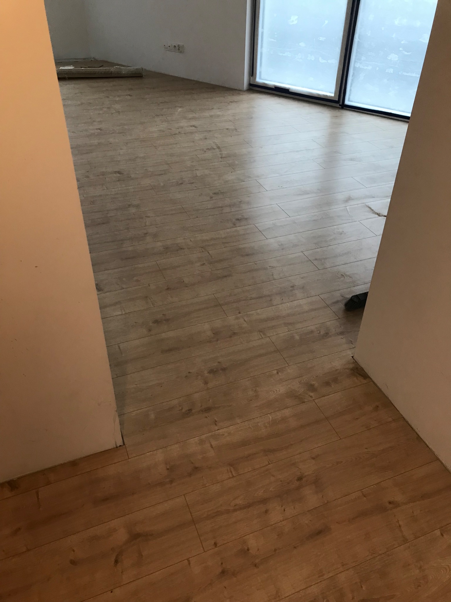 Widok przez futrynę drzwi na nowo ułożoną podłogę z paneli imitujących drewno, z widocznym oknem w tle i zwiniętym dywanem w rogu.