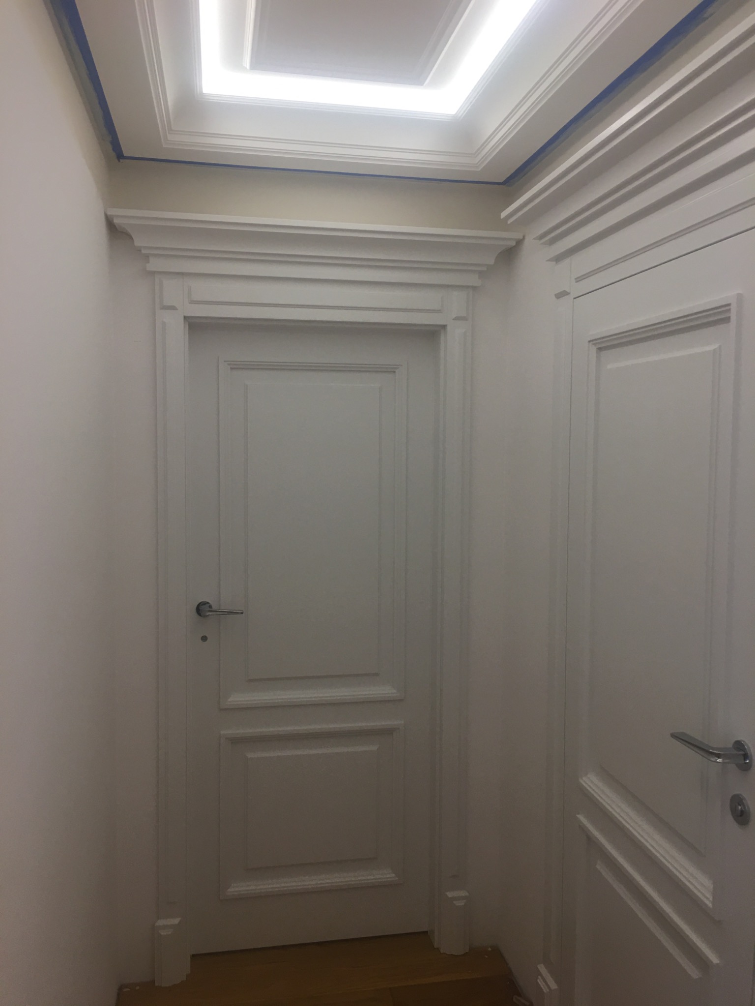 Dwa białe, klasyczne drzwi wewnętrzne z dekoracyjnymi listwami i srebrnymi klamkami, widoczne w korytarzu z drewnianą podłogą i sufitem z oświetleniem, zabezpieczonym niebieską taśmą malarską.