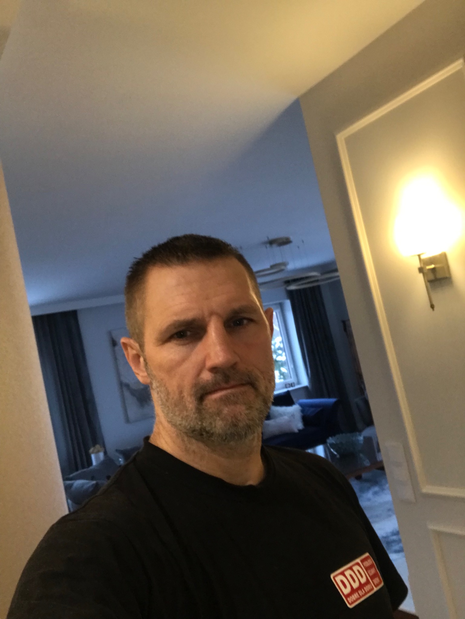 Mężczyzna w czarnej koszulce z logo firmy DDD na tle wnętrza mieszkania z widoczną lampą ścienną i fragmentem salonu.