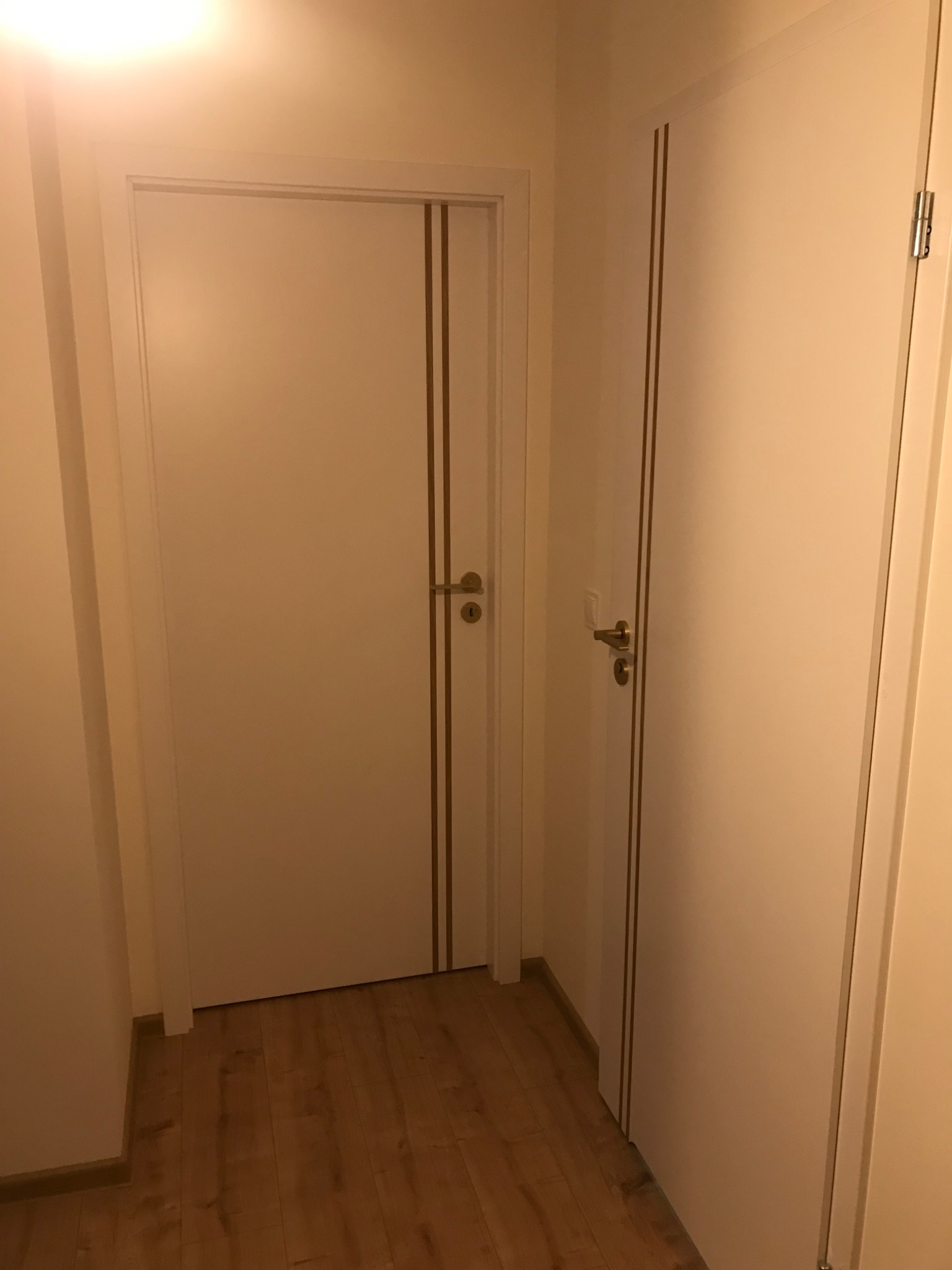 Dwie białe drzwi wewnętrzne z delikatnymi złotymi paskami, zamontowane w korytarzu z parkietem, widoczne klamki i zawiasy.