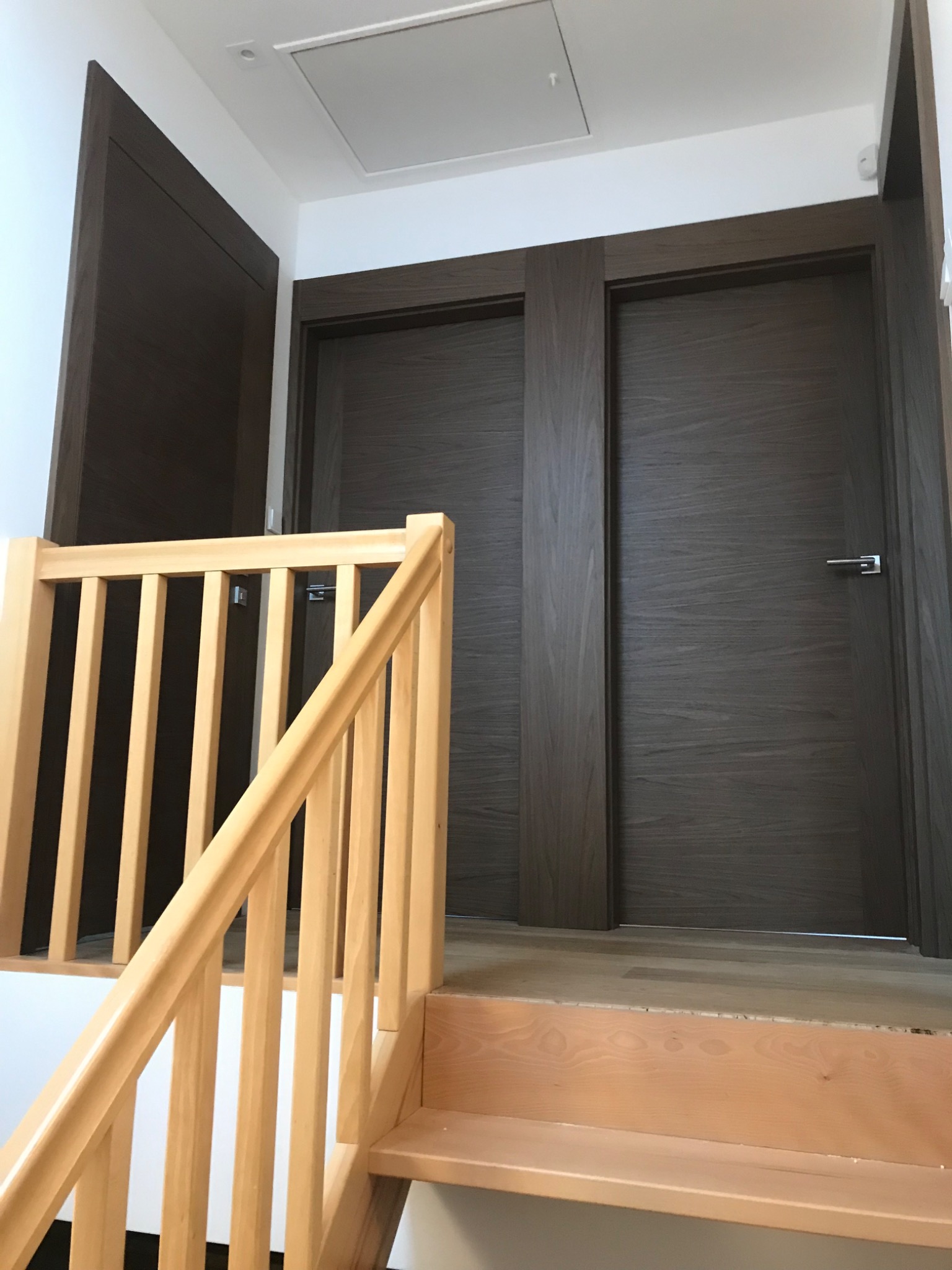 Dwa ciemne, drewniane drzwi z klamkami, widoczne zza jasnych, drewnianych schodów z barierką w nowym domu.