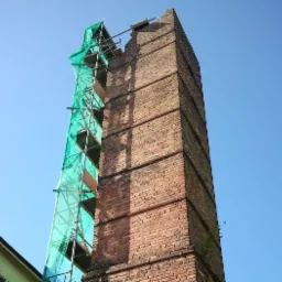 Wysoki, ceglany komin przemysłowy częściowo zrujnowany, z fragmentami rusztowania osłoniętego zieloną siatką, na tle błękitnego nieba.