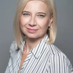 Portret uśmiechniętej kobiety z blond włosami do ramion, ubrana w koszulę w pionowe paski na szarym tle.