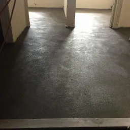 Świeżo wylana, gładka posadzka jastrychowa z widocznymi filarami i przejściami do innych pomieszczeń, w surowym stanie wykończenia.