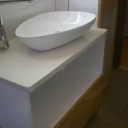 Owalna, biała umywalka nablatowa na białym blacie z drewnianym frontem szuflady, minimalistyczna łazienka z szarymi płytkami na ścianie.