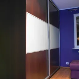 Wysoka szafa przesuwna z panelami z ciemnego drewna i białego szkła, widoczna na tle fioletowej ściany i drewnianej podłogi.