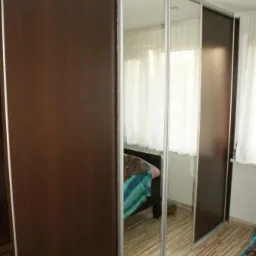 Duża szafa przesuwna z lustrami i ciemnobrązowymi panelami, odbijająca fragment sypialni z łóżkiem i oknem.