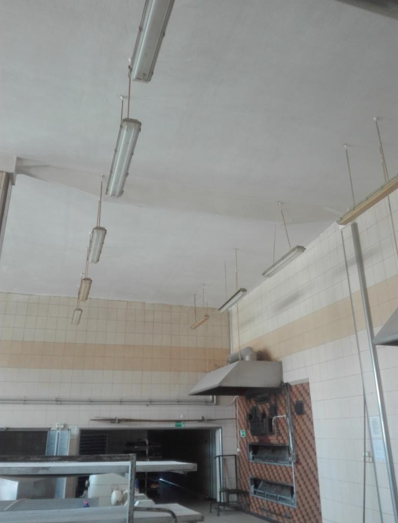 Wnętrze przemysłowej kuchni z białymi kafelkami na ścianach, widoczne lampy jarzeniowe pod sufitem i metalowy okap nad piecem.