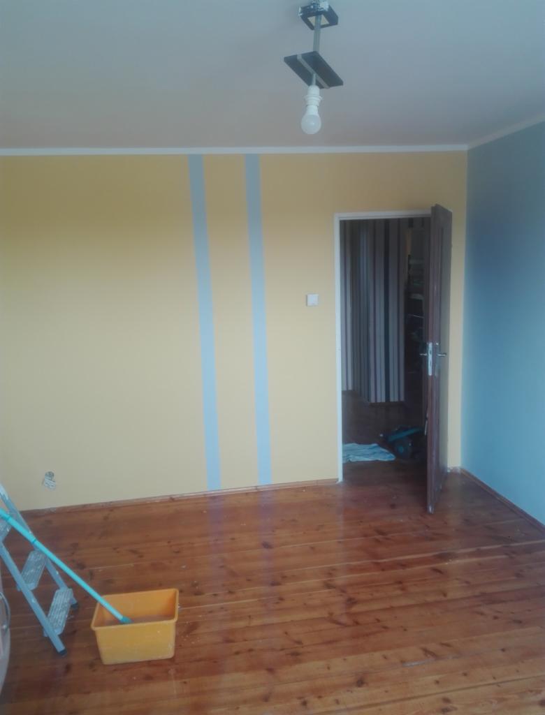 Pokój po malowaniu ścian na żółto i niebiesko z pionowymi pasami, widoczna drabina malarska i kuweta z farbą na lakierowanej, drewnianej podłodze.