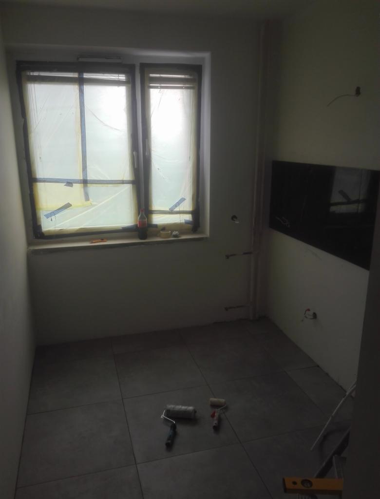 Wnętrze pokoju w trakcie prac wykończeniowych: zabezpieczone okno folią, położona podłoga z szarych płytek, wałki malarskie na podłodze, poziomica przy ścianie.