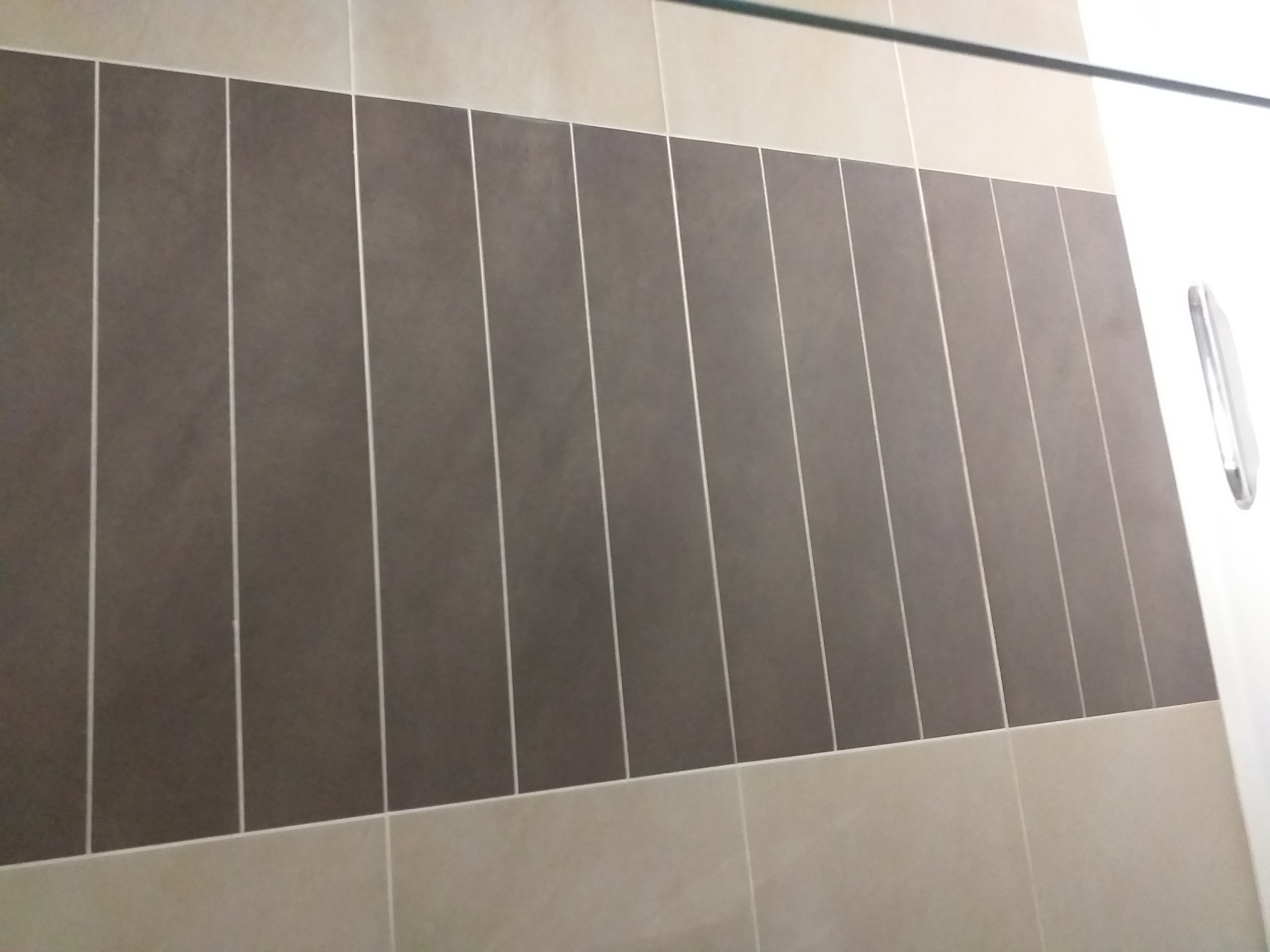 Fragment ściany wyłożonej dwukolorowymi, pionowymi płytkami ceramicznymi w odcieniach beżu i szarości, z widoczną fugą i fragmentem kabiny prysznicowej.