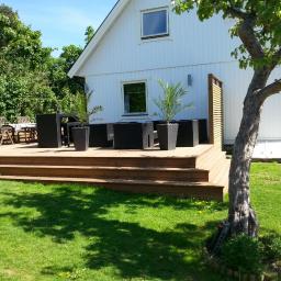 AM Firma - Drewniany taras z niskimi schodami przed białym domem, wyposażony w czarne meble ogrodowe i donice z palmami, częściowo zacieniony przez drzewo.