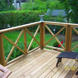AM Firma - Wykończony taras z drewnianą balustradą w kształcie krzyża, widok na zielony trawnik i krzewy, z leżakiem i fragmentem stołu, lampa solarna na słupku balustrady.