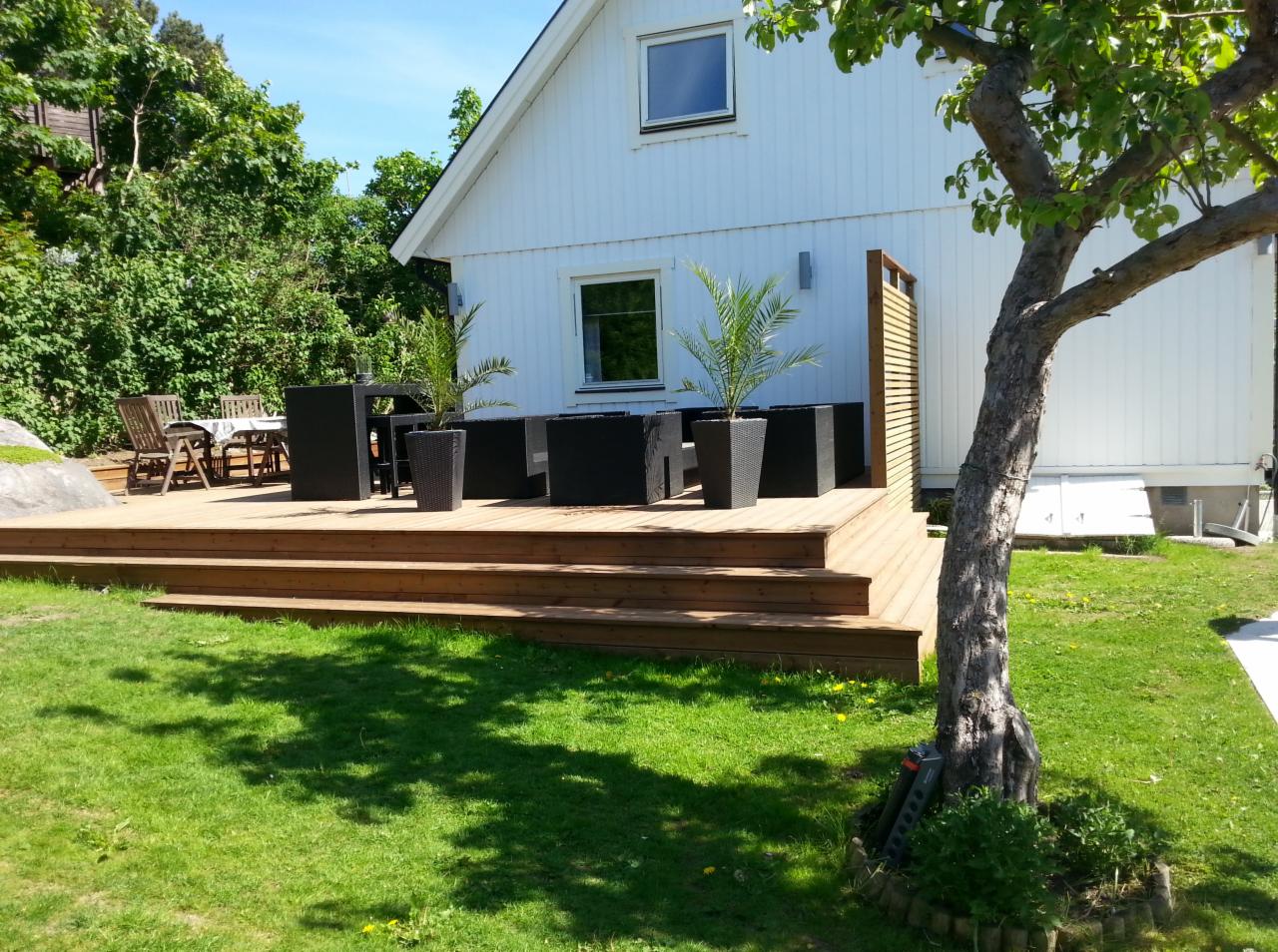 Drewniany taras z niskimi schodami przed białym domem, wyposażony w czarne meble ogrodowe i donice z palmami, częściowo zacieniony przez drzewo.