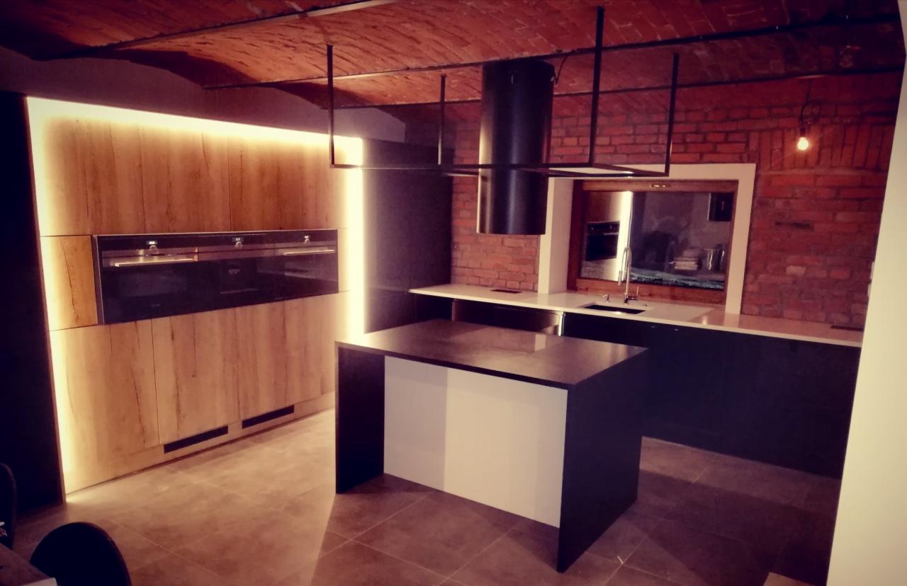 Nowoczesna kuchnia z wyspą, drewnianymi frontami szafek podświetlonymi ledami, ceglanym sufitem i ścianą, czarnym okapem oraz elementami w kolorze antracytu.