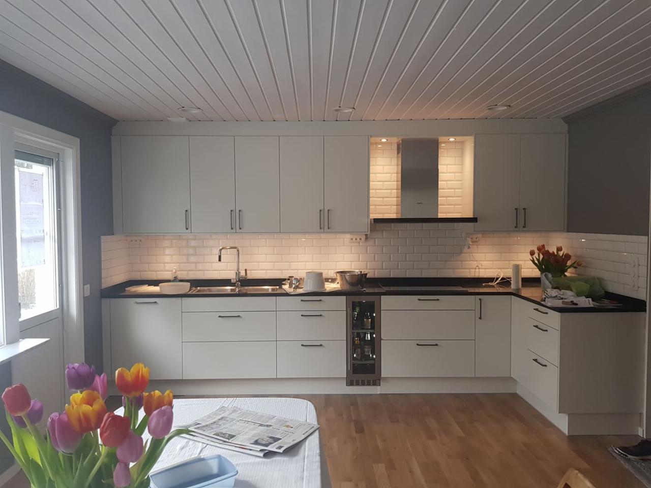 Jasna kuchnia z białymi szafkami i czarnym blatem w Hucisku, wykończona białymi płytkami typu metro, z widoczną zabudowaną lodówką na napoje.