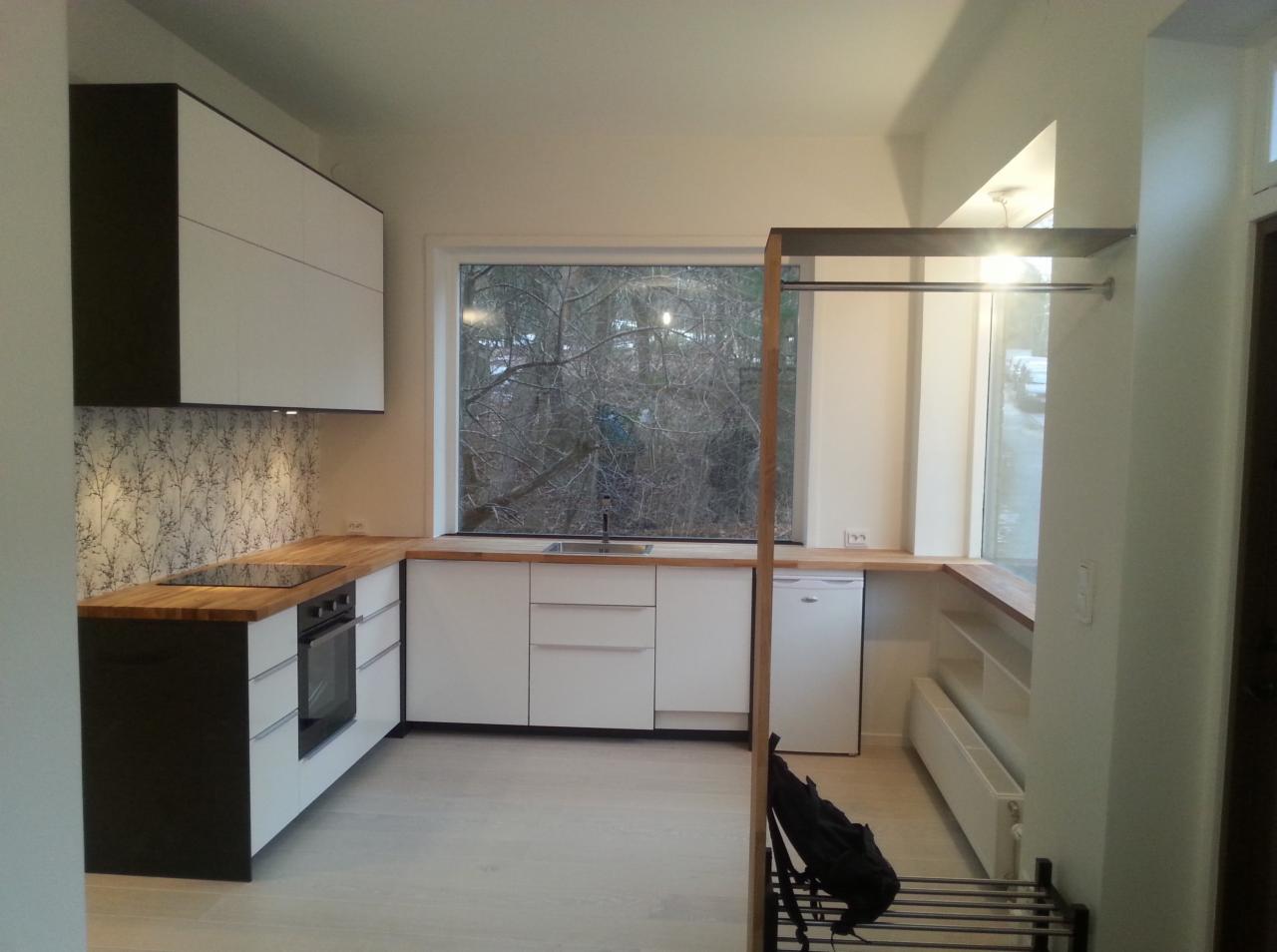 Nowoczesna kuchnia z białymi frontami szafek, drewnianym blatem i tapetą w delikatny wzór gałązek, widok z okna na drzewa bez liści, fragment garderoby z metalowym wieszakiem.