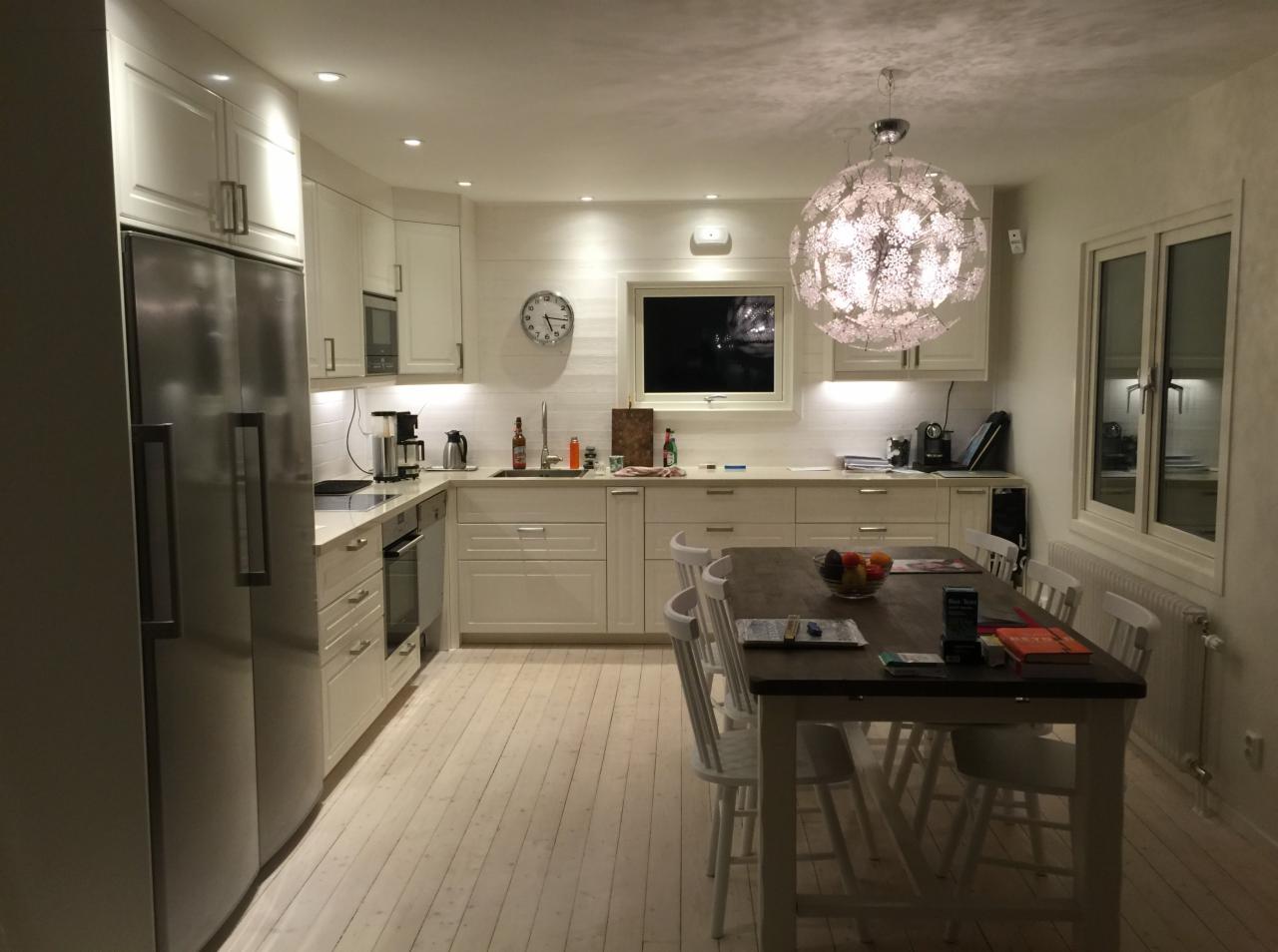 Jasna, przestronna kuchnia z białymi meblami, drewnianym stołem i designerską lampą w kształcie kuli, widok na okno i sprzęty AGD.