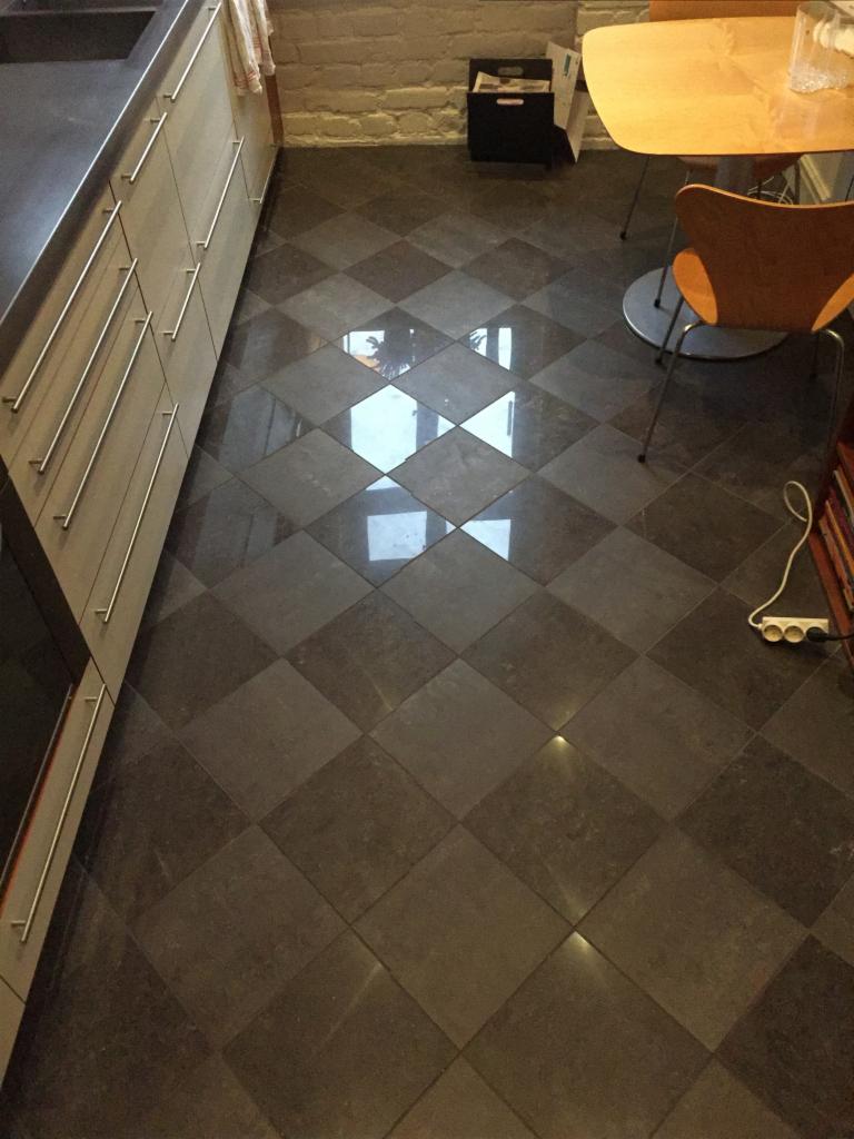 Ukończona podłoga w kuchni z ciemnych, kwadratowych płytek ułożonych w karo, widoczny fragment zabudowy kuchennej z szufladami z metalowymi uchwytami, stół z krzesłami i fragment ściany z cegły.