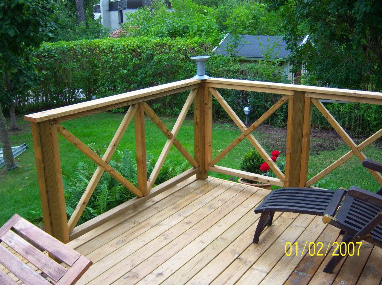 Wykończony taras z drewnianą balustradą w kształcie krzyża, widok na zielony trawnik i krzewy, z leżakiem i fragmentem stołu, lampa solarna na słupku balustrady.