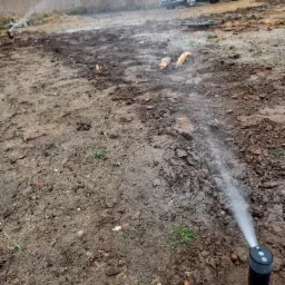 Instalacja nawadniania trawnika w trakcie pracy, widoczny spryskiwacz w ziemi oraz mokra, błotnista gleba, w tle zaparkowany samochód osobowy.