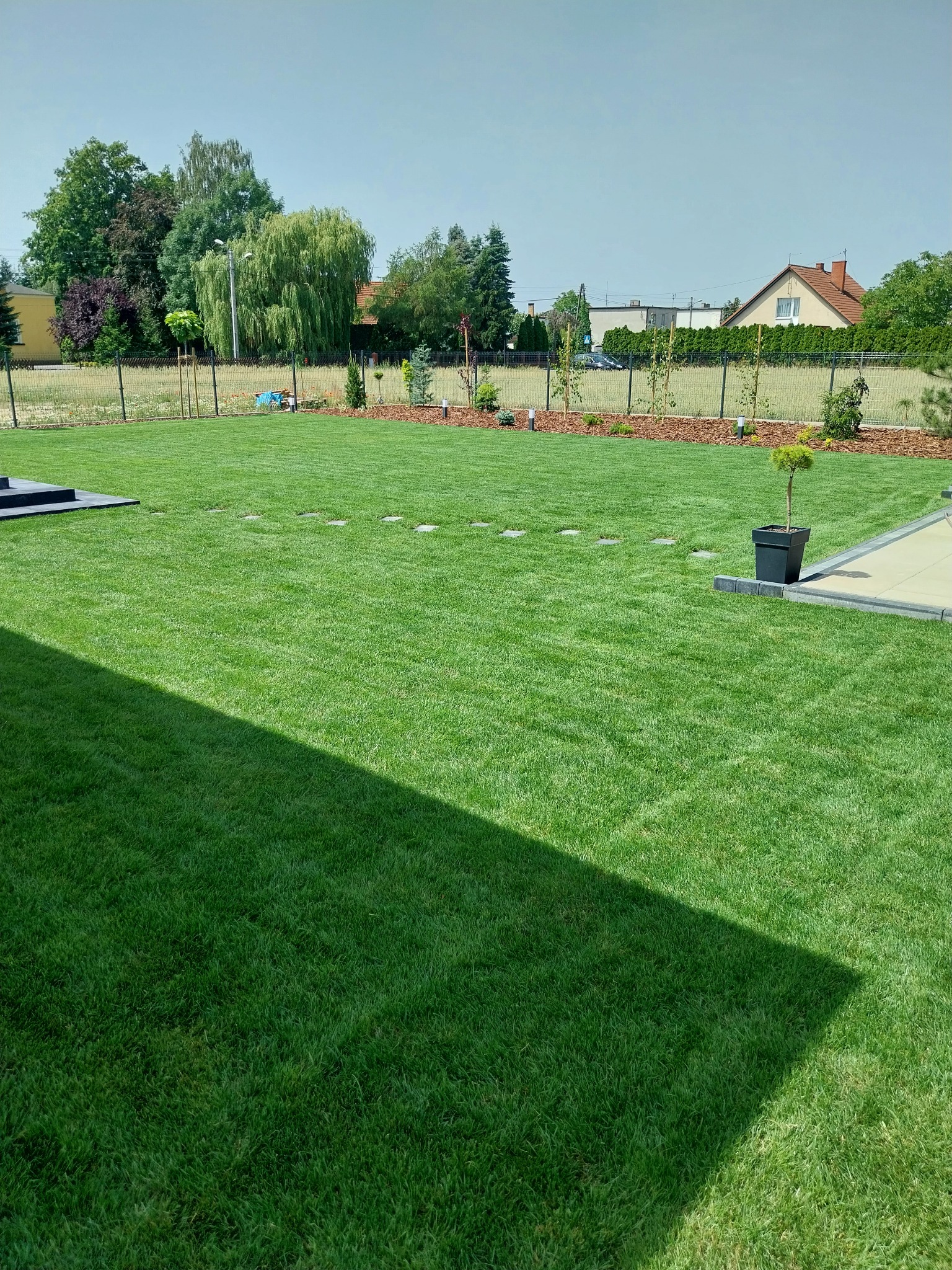 Rozległy, równo przystrzyżony trawnik z zieloną trawą, ozdobne drzewka wzdłuż ogrodzenia, ścieżka z kamiennych płyt i cień na pierwszym planie.