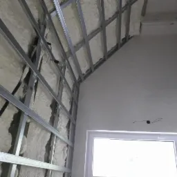 Poddasze w trakcie izolacji pianą PUR, widoczne metalowe profile konstrukcyjne, okno i fragment ściany z instalacją elektryczną.