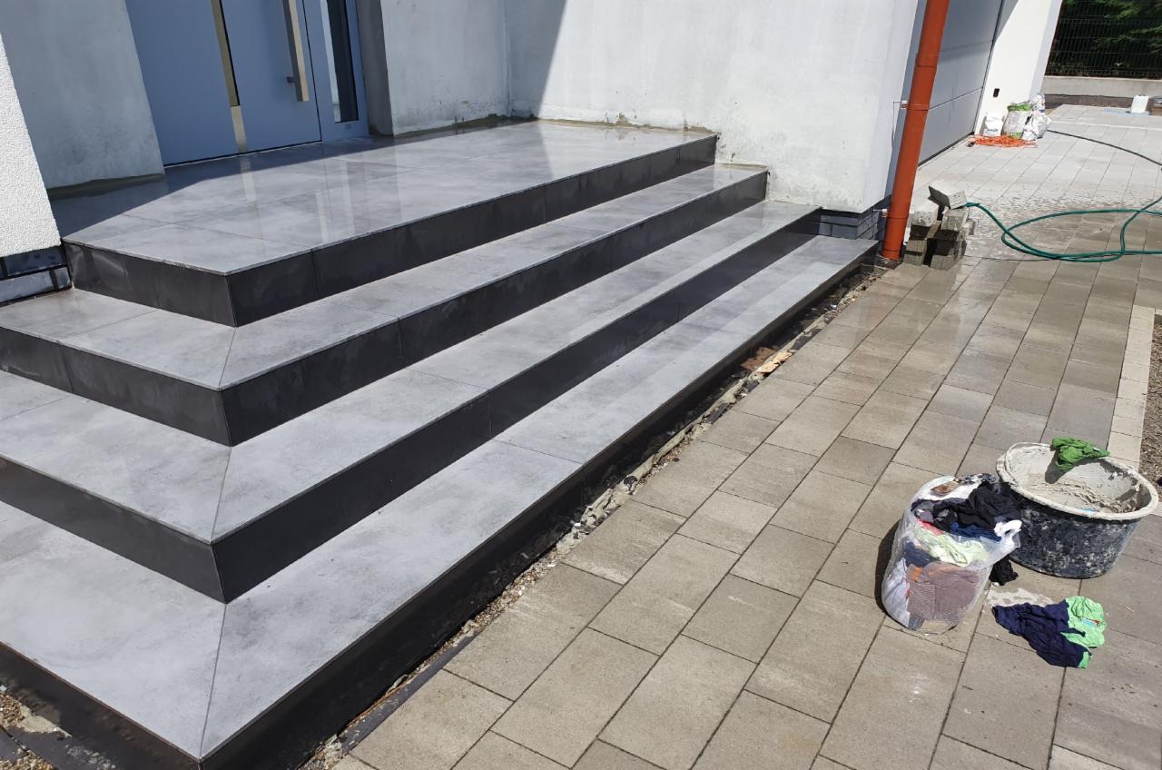 Wykończone zewnętrzne schody wejściowe do domu, pokryte szarymi płytkami z czarnymi krawędziami, obok widoczne wiadro i worek z narzędziami.
