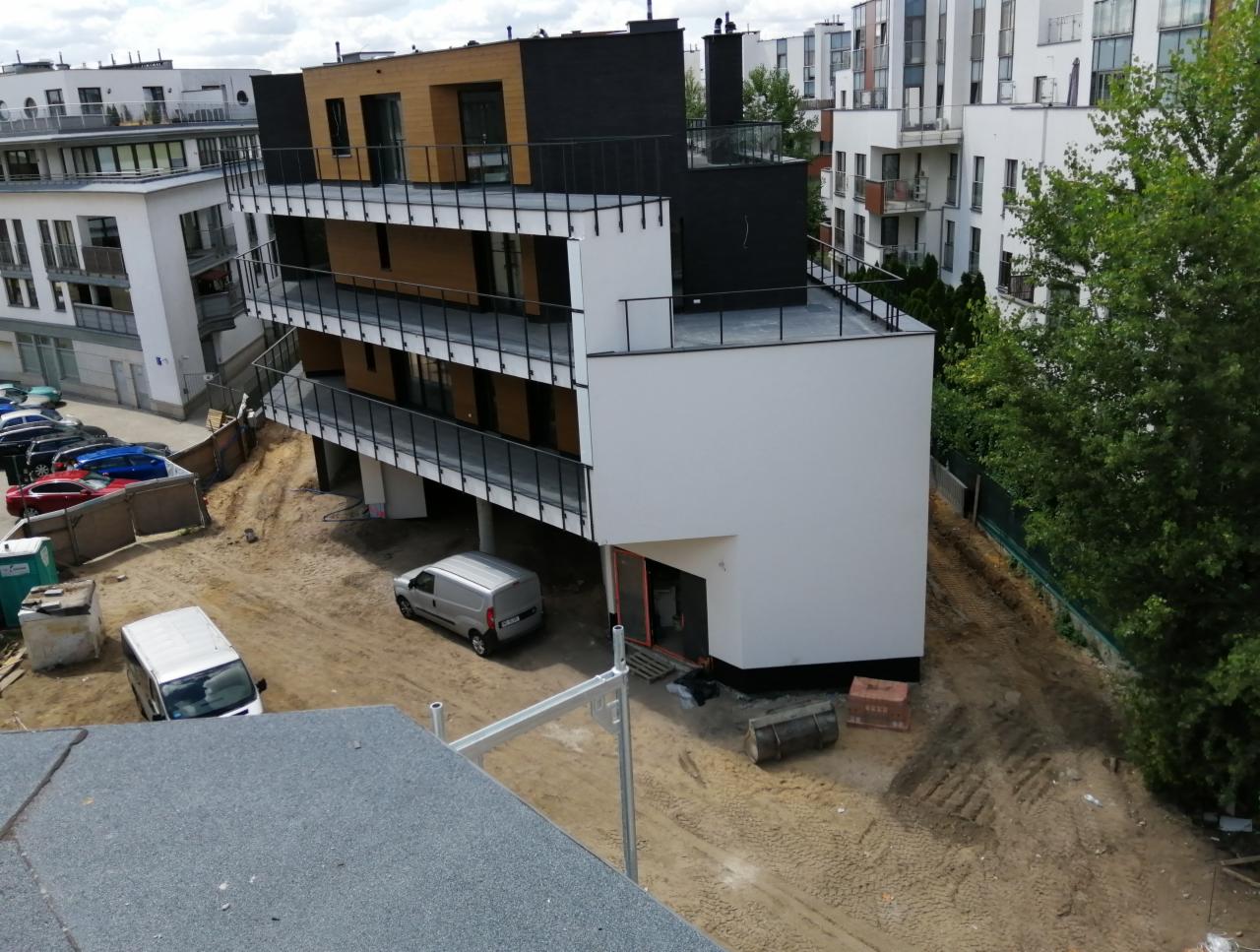 Nowoczesny budynek wielorodzinny z białymi ścianami i balkonami z czarnymi balustradami, widok z góry na plac budowy przed budynkiem, zaparkowane samochody i teren przygotowany pod zagospodarowanie.