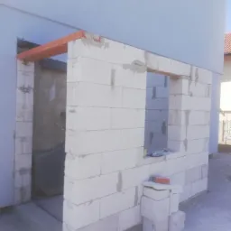 Wznoszenie ściany z białych bloczków gazobetonowych z widocznym otworem okiennym i nadprożem z czerwonej cegły, etap budowy domu.