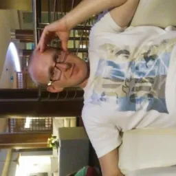 Mężczyzna w okularach, leżący na jasnej kanapie, ubrany w białą koszulkę z nadrukiem, z ręką na głowie, w tle wnętrze budynku z kolumnami i oknami.