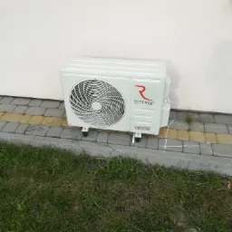 Biała jednostka zewnętrzna klimatyzatora Rotenso zamontowana na elewacji domu, widoczne ślady po ołówku na kostce brukowej, trawa w tle.