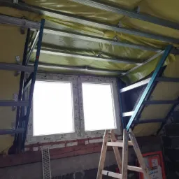 Montaż stelaża pod sufit podwieszany na poddaszu, widoczne profile metalowe, izolacja żółtą folią, okno dachowe i drabina. W tle ściana z cegły i karton z napisem JomoTech.