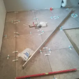 Podłoga w trakcie układania płytek z wzorem, użyte poziomice, kliny i system poziomowania, widoczny worek z klejem i fragment instalacji wodnej.